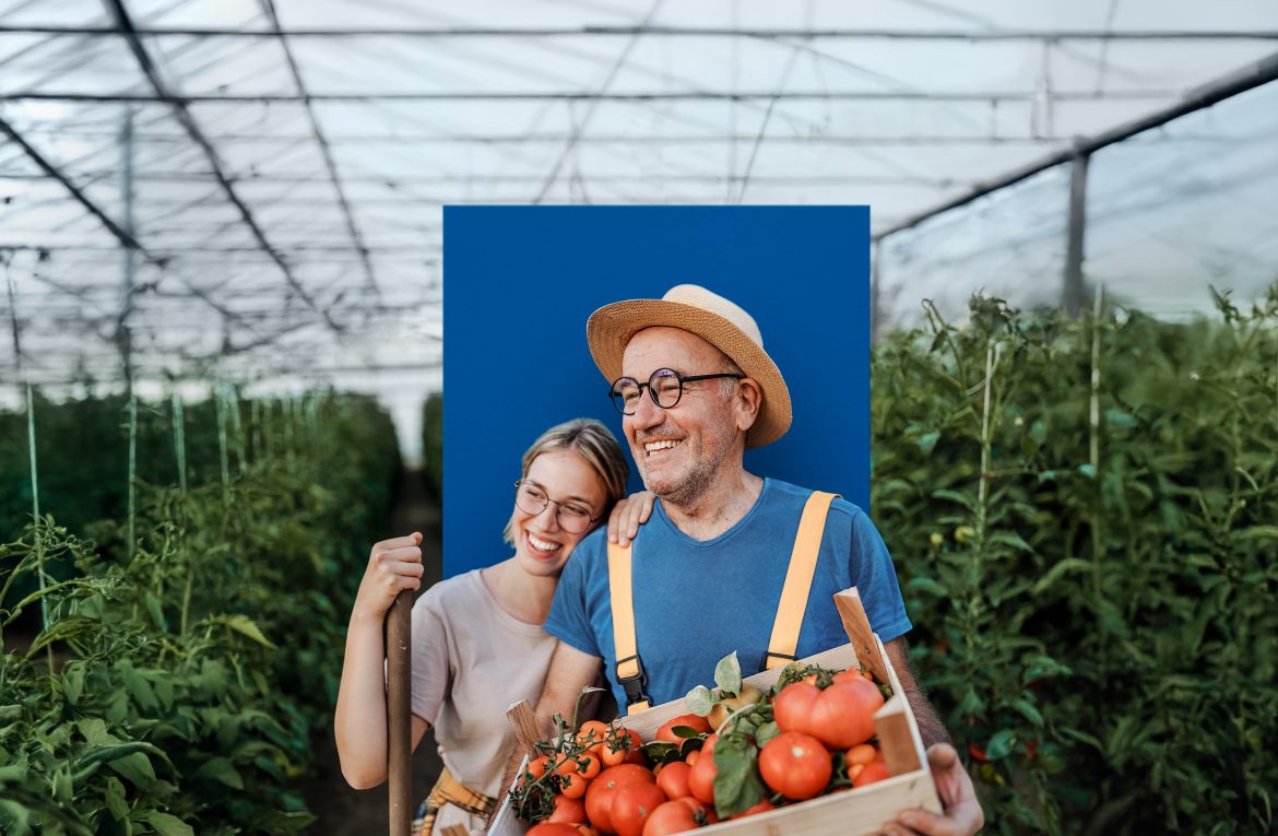 Ein Vater steht in einem Gewächshaus und hält einen Korb Gemüse. Seine Tochter lehnt ihren Kopf lachend an seine Schulter.
