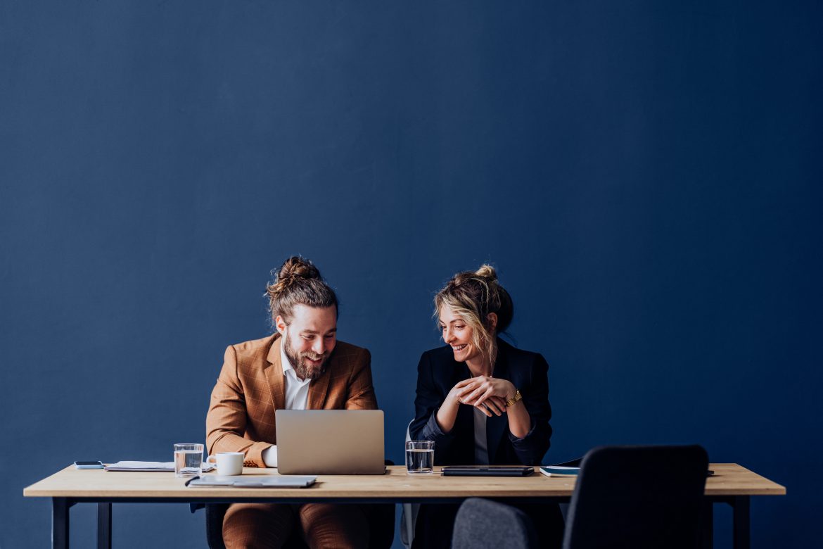 zwei kollegen sitzen vor blauem hintergrund lachend vor einem laptop