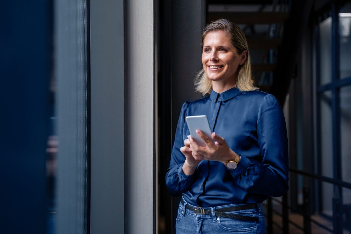 Happy businesswoman standing with smart phone in office corridor